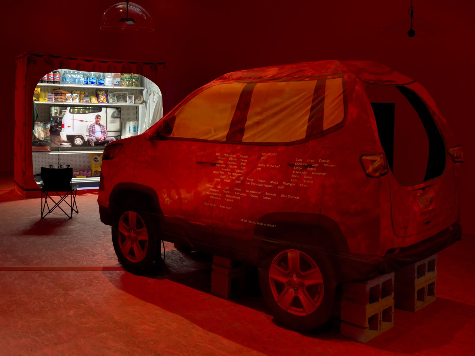 An art installation featuring a soft material vehicle, a camping chair, and a projector playing a film.