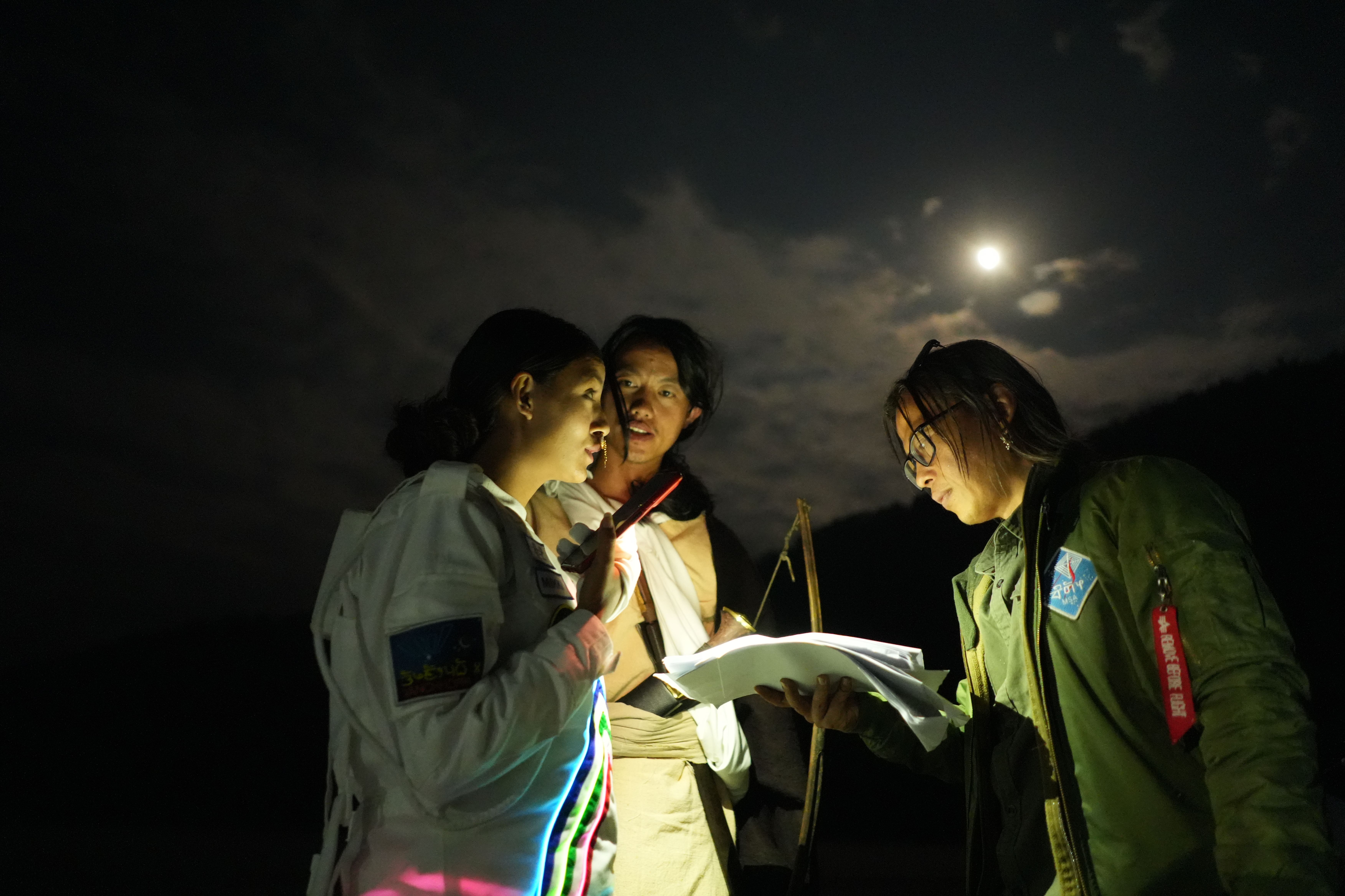 Three individuals stand outdoor at night, engrossed in reviewing papers. One of them holds a lamp emitting a warm glow, il...