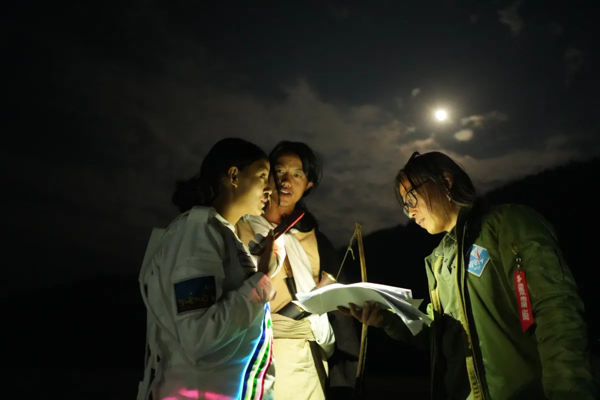Three individuals stand outdoor at night, engrossed in reviewing papers. One of them holds a lamp emitting a warm glow, illuminating their focused faces and the papers in hand.
