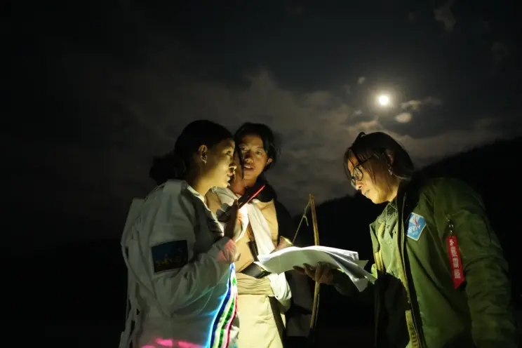 Three individuals stand outdoor at night, engrossed in reviewing papers. One of them holds a lamp emitting a warm glow, il...
