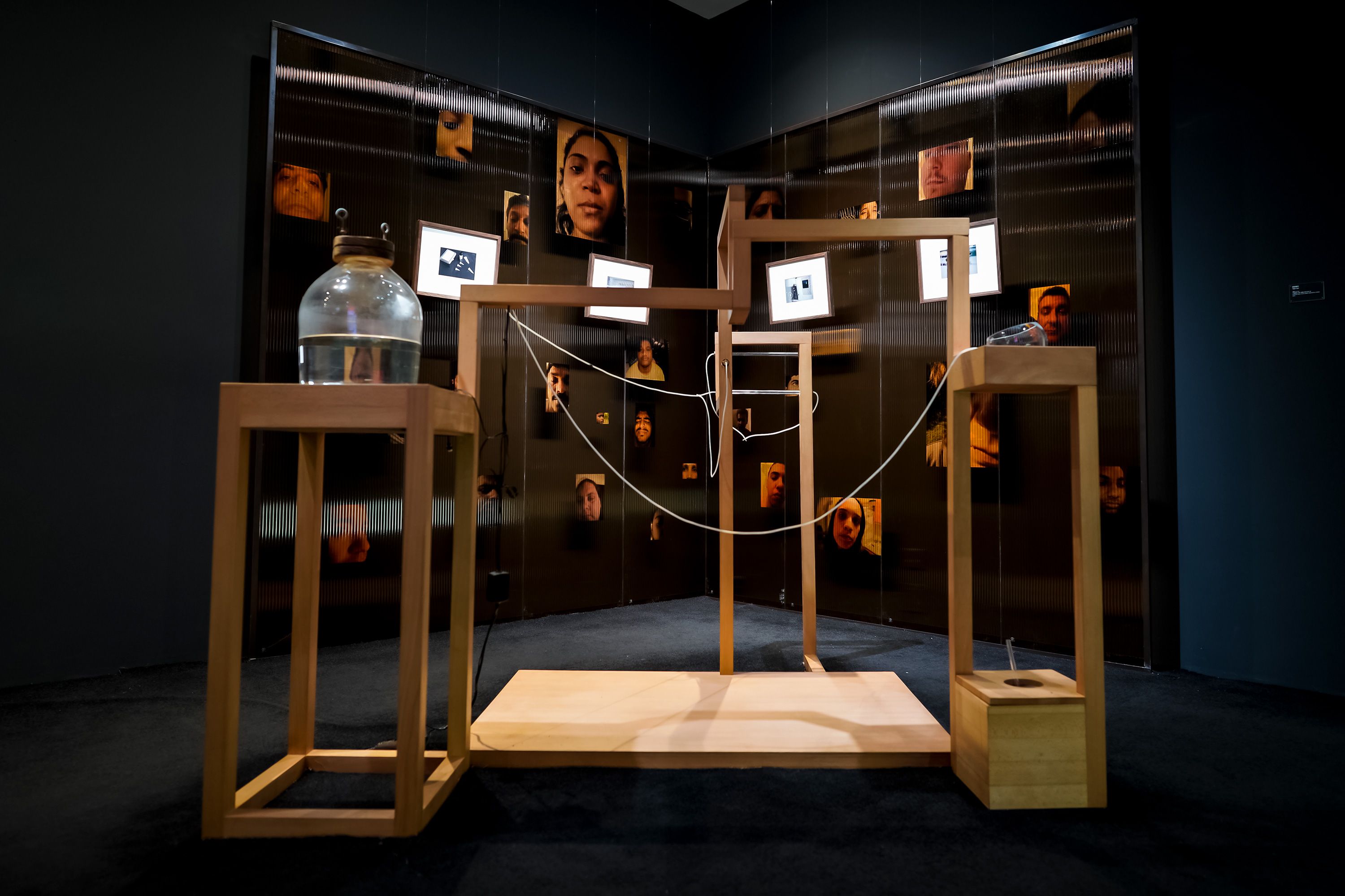 An art installation featuring a wooden construction adorned with hanging white wires, a jar filled with water, and sepia p...