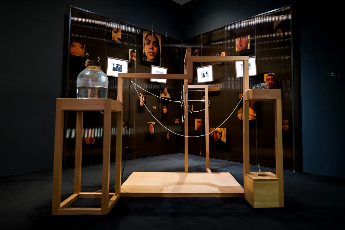 An art installation featuring a wooden construction adorned with hanging white wires, a jar filled with water, and sepia photos of people's faces in the background.