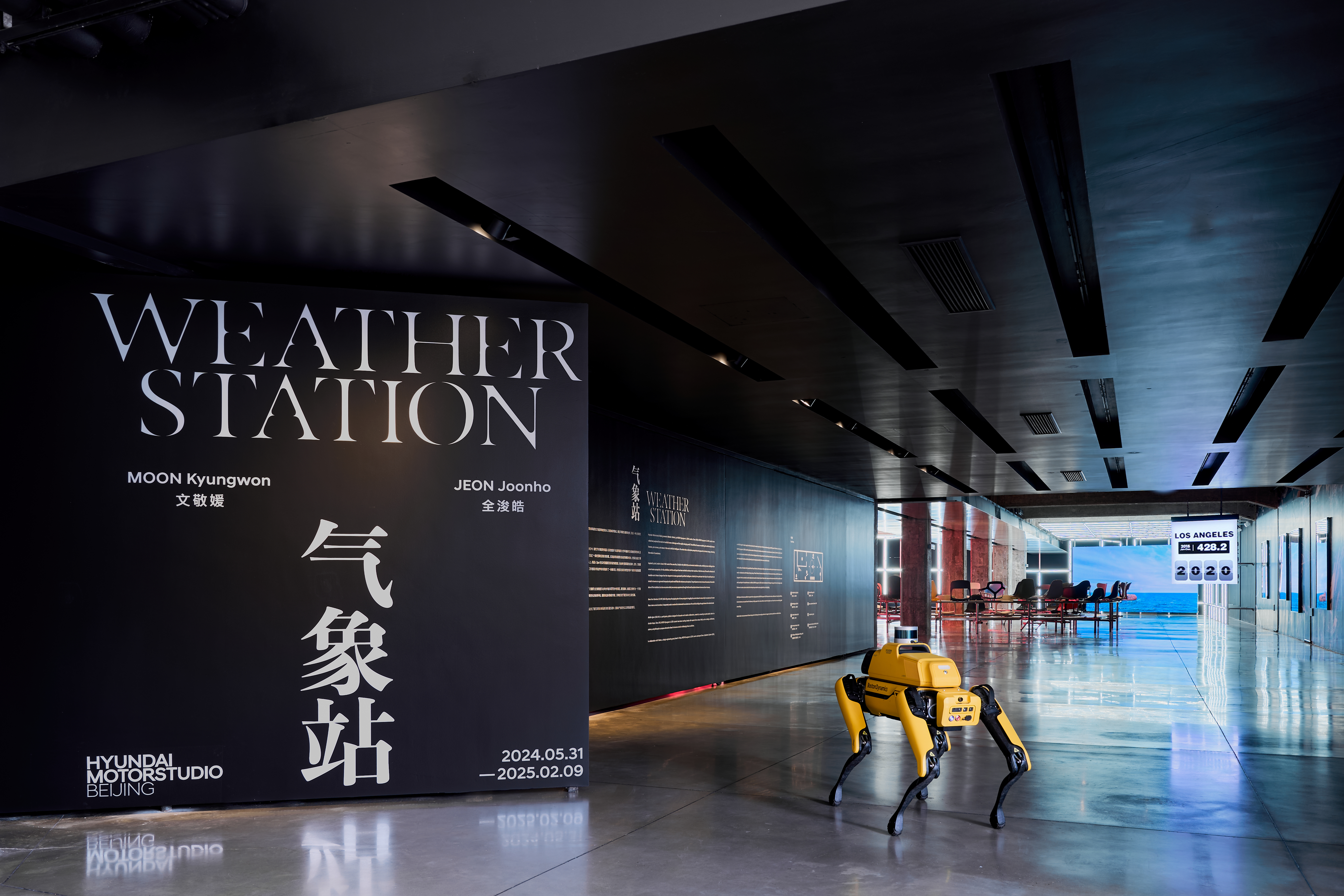 A modern, sleek exhibition space with a large black wall displaying "Weather Station" and the names "Moon Kyungwon" and "J...