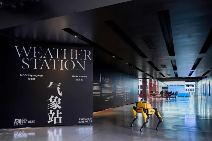 A modern, sleek exhibition space with a large black wall displaying "Weather Station" and the names "Moon Kyungwon" and "J...
