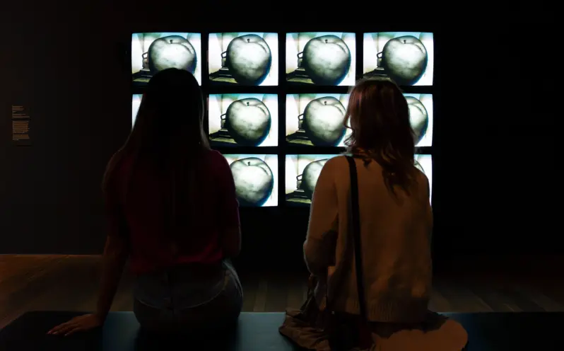 Two women sit in a dark gallery facing a video installation composed of twelve monitors arranged in a grid. Each screen di...