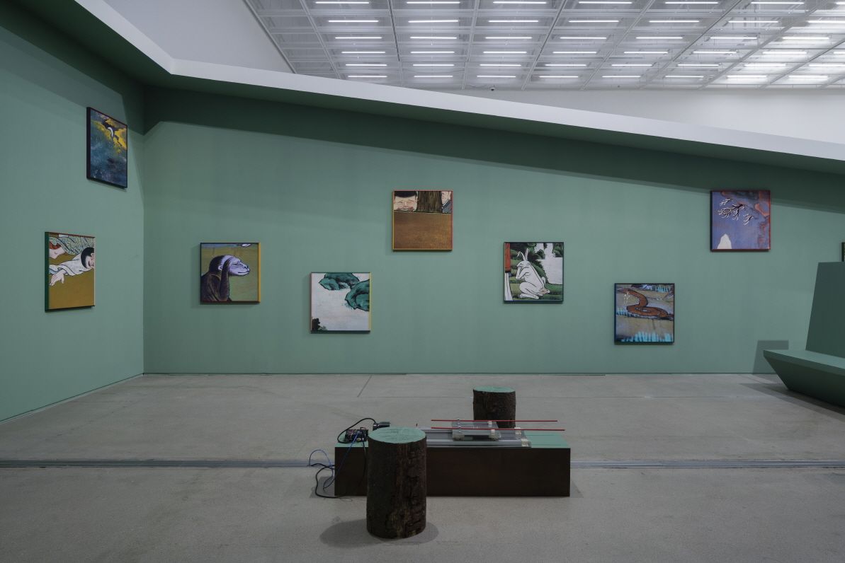 A bright gallery room featuring green walls adorned with framed artworks. In the center of the room, an art installation c...