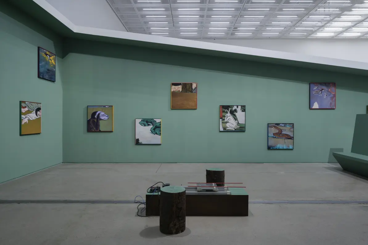 A bright gallery room featuring green walls adorned with framed artworks. In the center of the room, an art installation consisting of two cylinders and a rectangular object with unidentified electronics on top is displayed. A green bench is also present in the room.