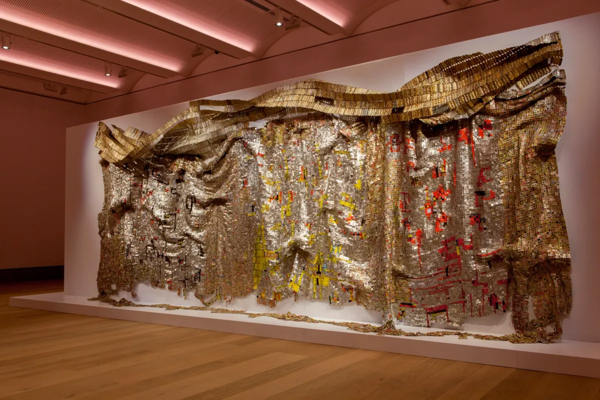 A large artwork by El Anatsui, made of aluminium and copper wire, showcasing intricate gold, brown, red and yellow patterns, displayed in a spacious gallery room.