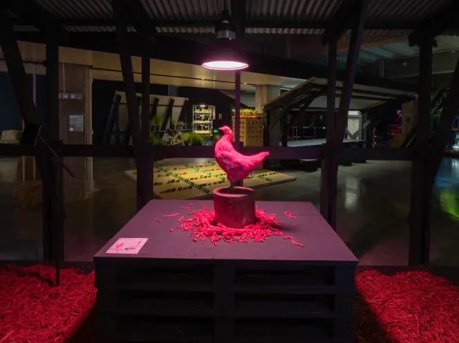 Pink bird sculpture on a pedestal with red shavings, illuminated by an overhead lamp.