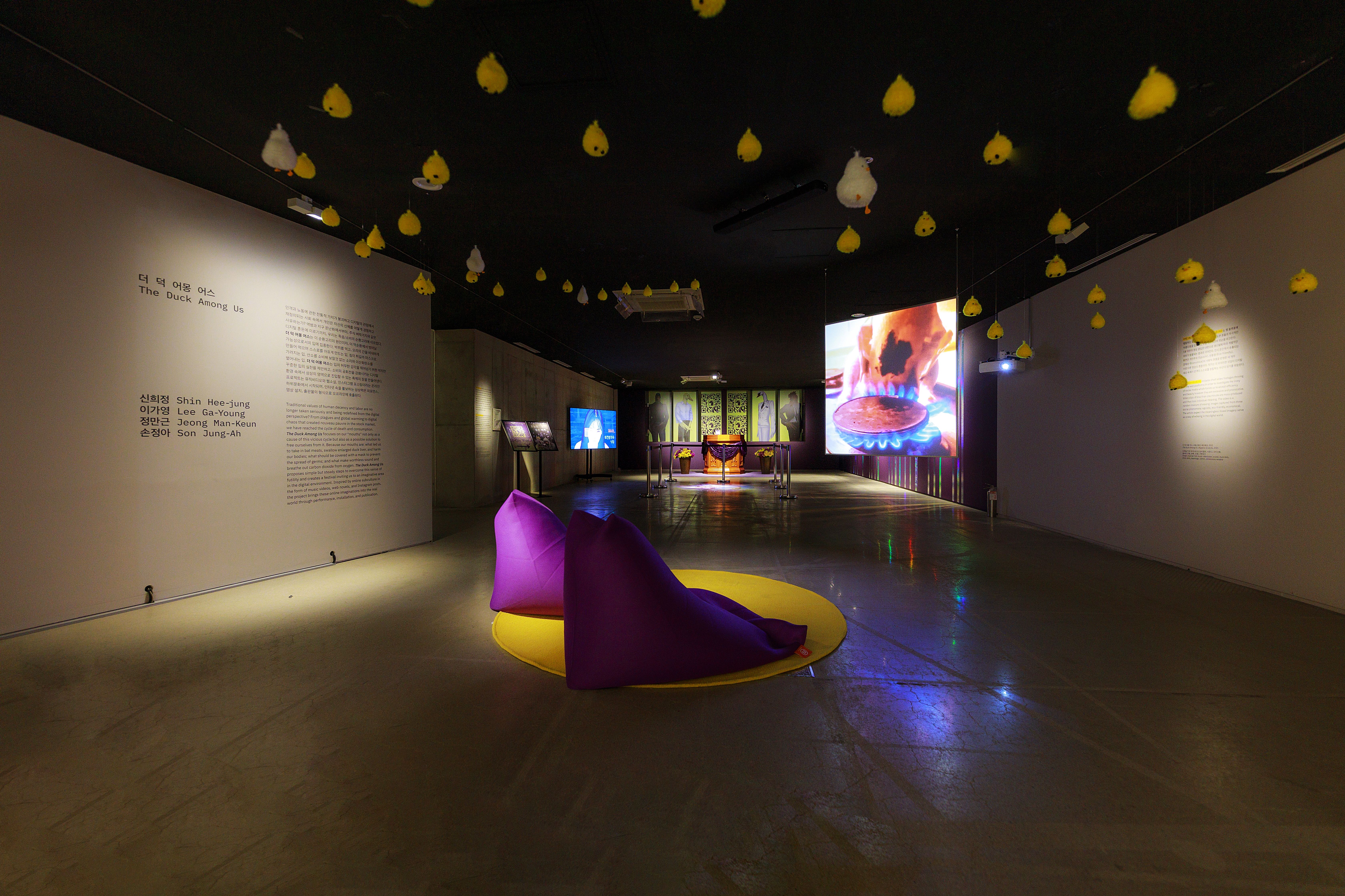 An art installation in a gallery space with white and yellow objects suspended from the ceiling. Two purple beanbags are p...