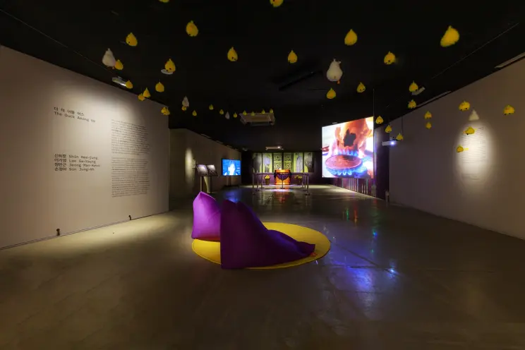 An art installation in a gallery space with white and yellow objects suspended from the ceiling. Two purple beanbags are p...