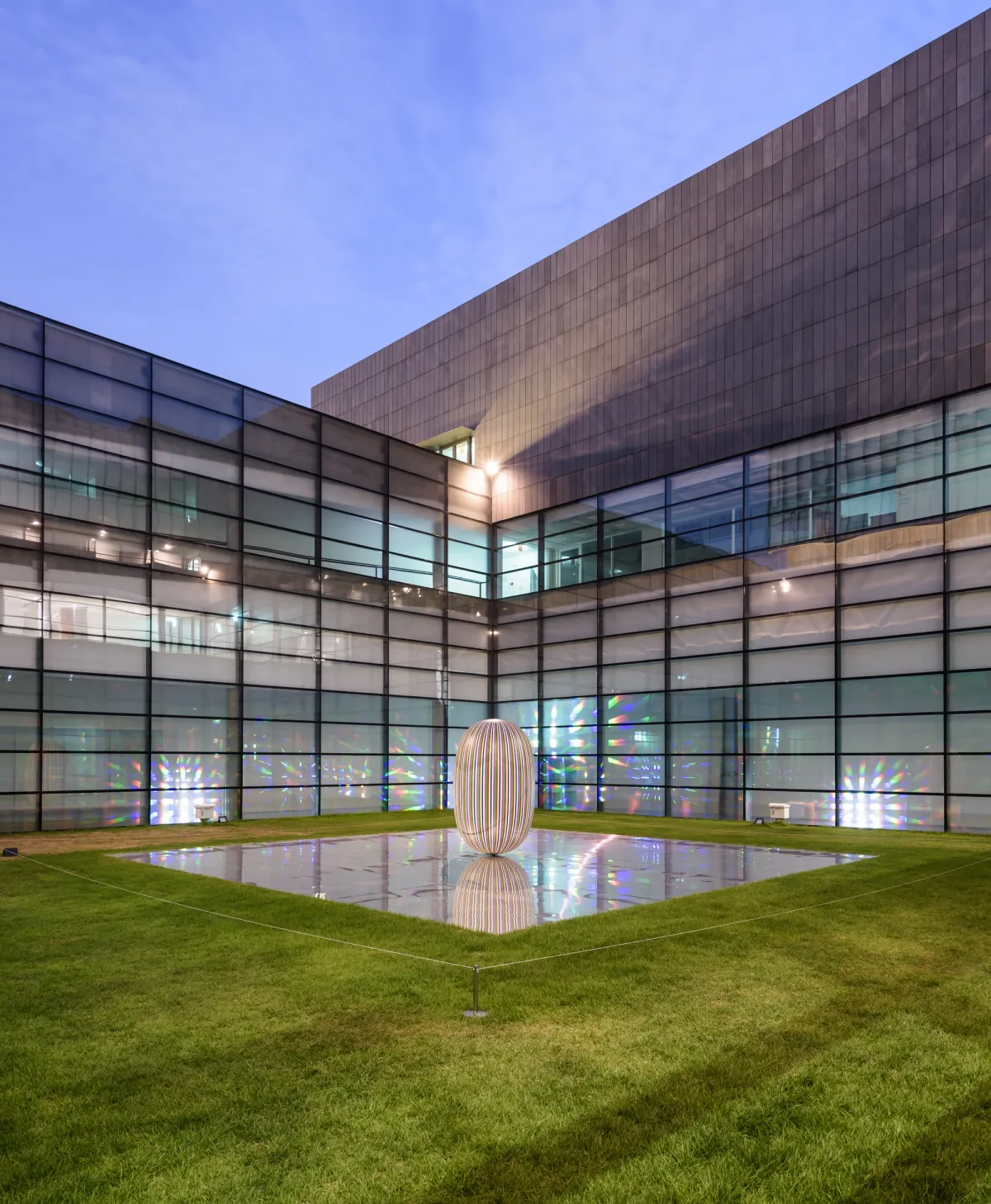 An elliptic sculpture situated on a mirrored rectangular surface in the backyard of a building with numerous windows. The sculpture's smooth, curved form contrasts with the building's angular architecture.