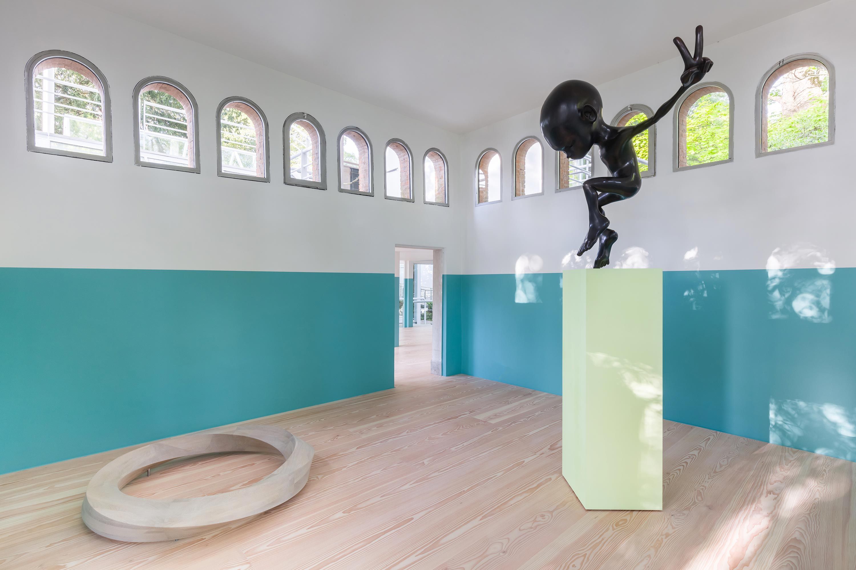 Sculpture of a black humanoid figure on a green pedestal in a room with teal and white walls, wooden flooring, arched wind...