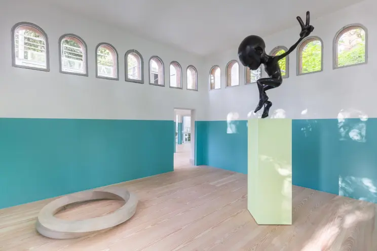 Sculpture of a black humanoid figure on a green pedestal in a room with teal and white walls, wooden flooring, arched wind...