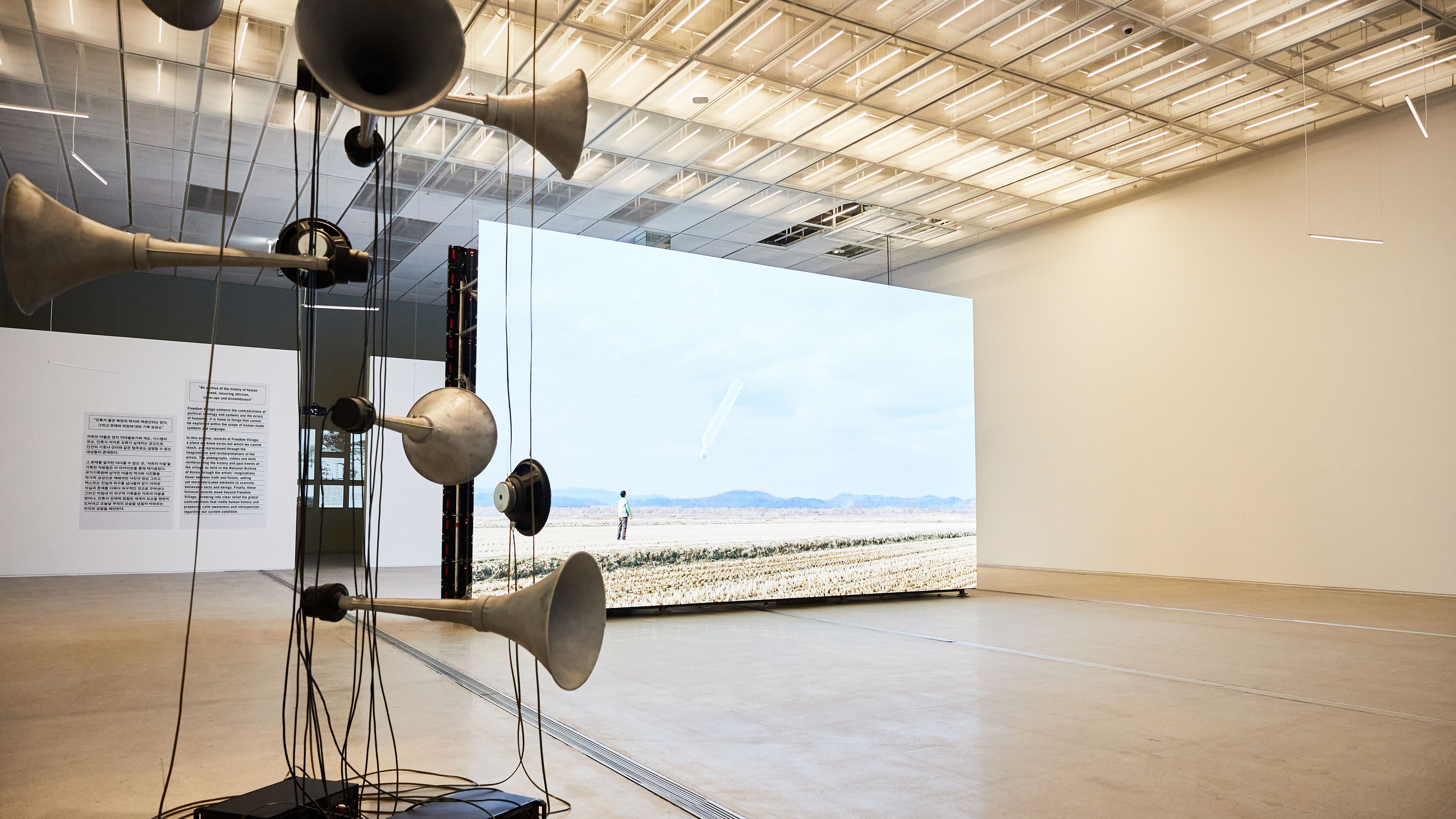 A large gallery space displays a life-sized photograph of a person standing in a field. An abstract sculpture, comprised o...