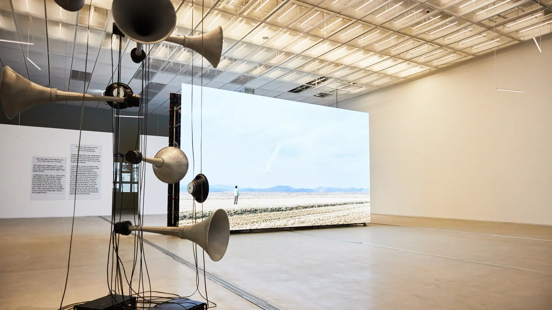 A large gallery space displays a life-sized photograph of a person standing in a field. An abstract sculpture, comprised o...