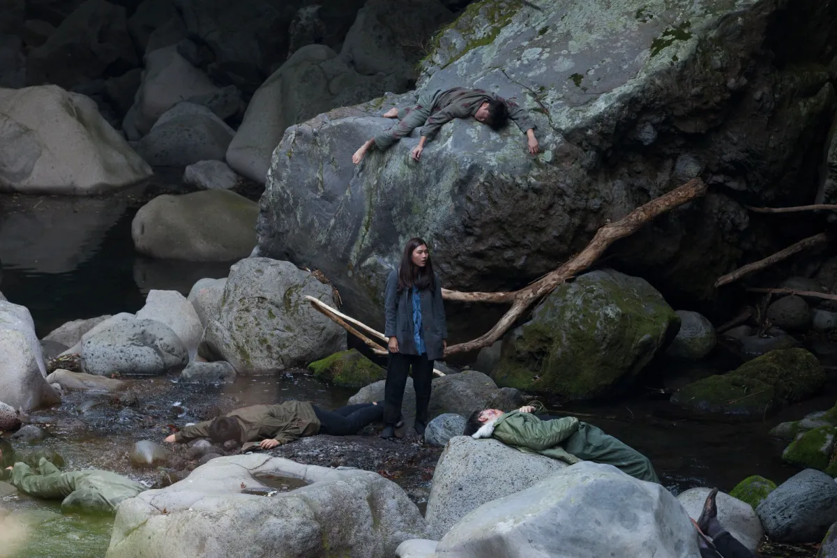 A person stands amidst massive boulders and a body of water, surrounded by figures lying motionless on the ground.