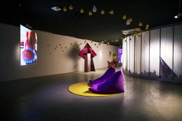 A gallery space featuring white and yellow duck and chick dolls hanging from the ceiling. Two purple beanbags are position...