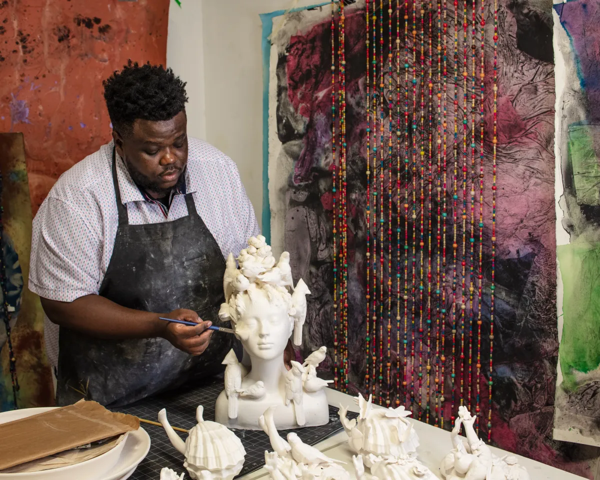 Morel Doucet is in his studio, diligently working on a sculpture featuring a person and birds. Beaded curtains and abstract art serve as a background.