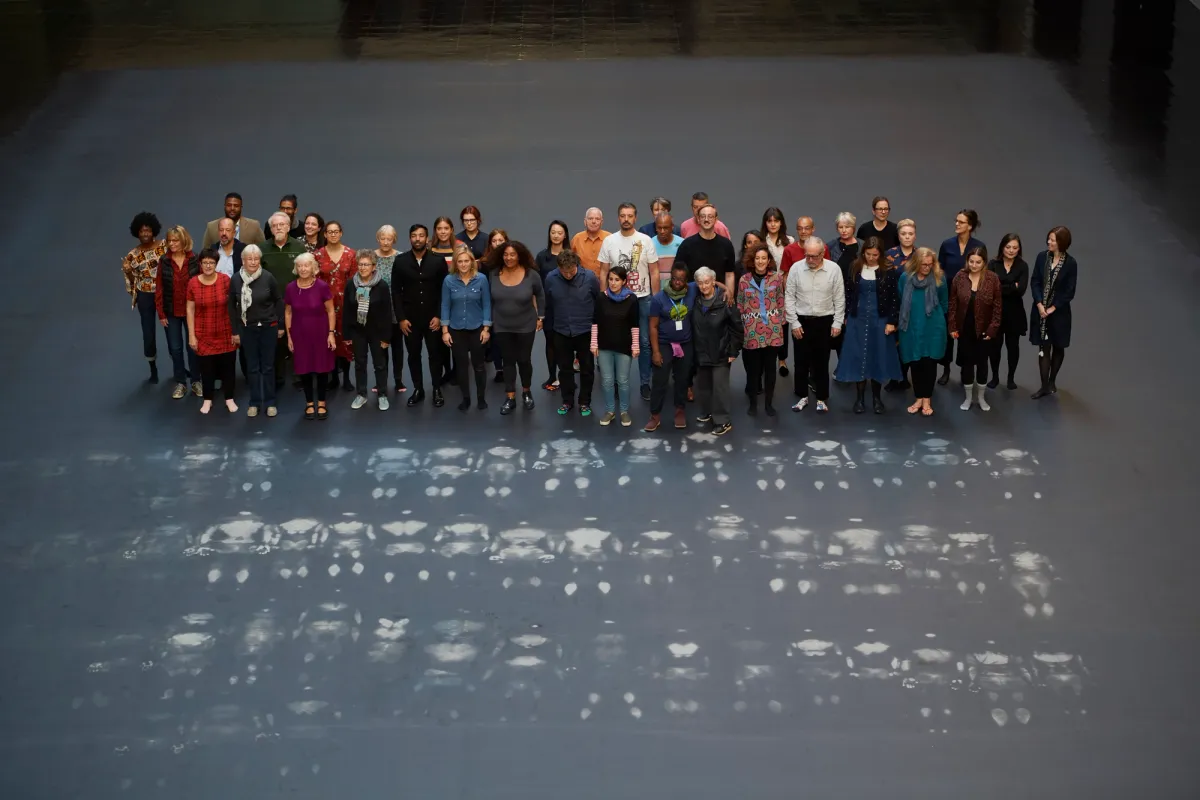 A crowd of people standing near three parallel rows of white outlines on a grey floor, marking the shapes of lying figures. The image captures a moment of contemplation as the crowd studies these traces.