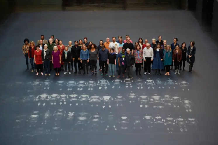 A crowd of people standing near three parallel rows of white outlines on a grey floor, marking the shapes of lying figures...