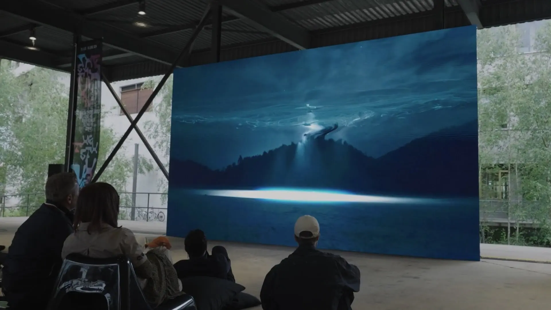 People watching a large outdoor screen displaying a blue mountainous landscape with a beam of light illuminating the ground.