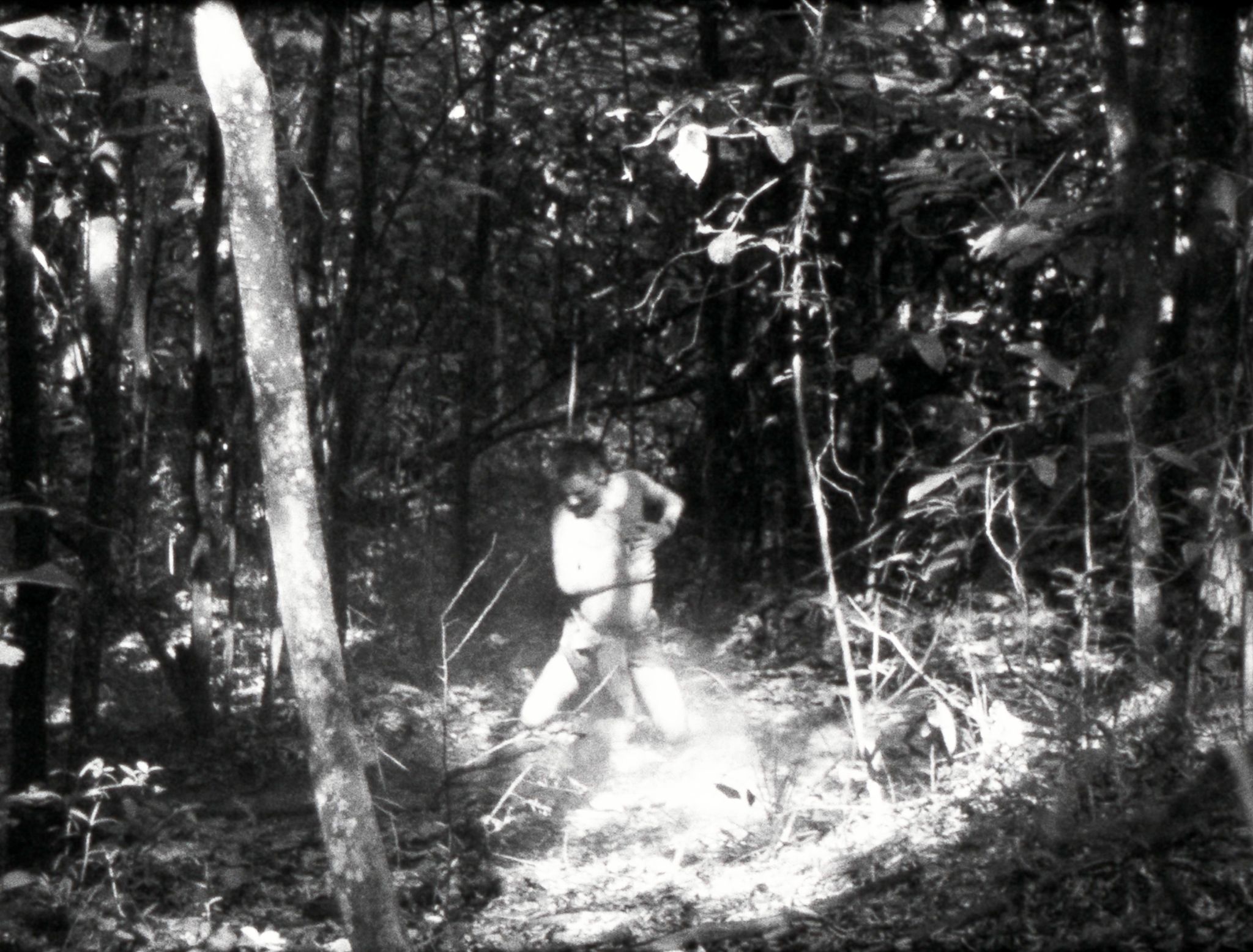 A black-and-white scene of a figure standing and moving through a sunlit forest clearing.