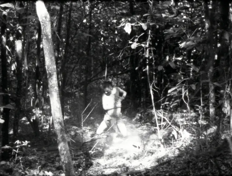 A black-and-white scene of a figure standing and moving through a sunlit forest clearing.