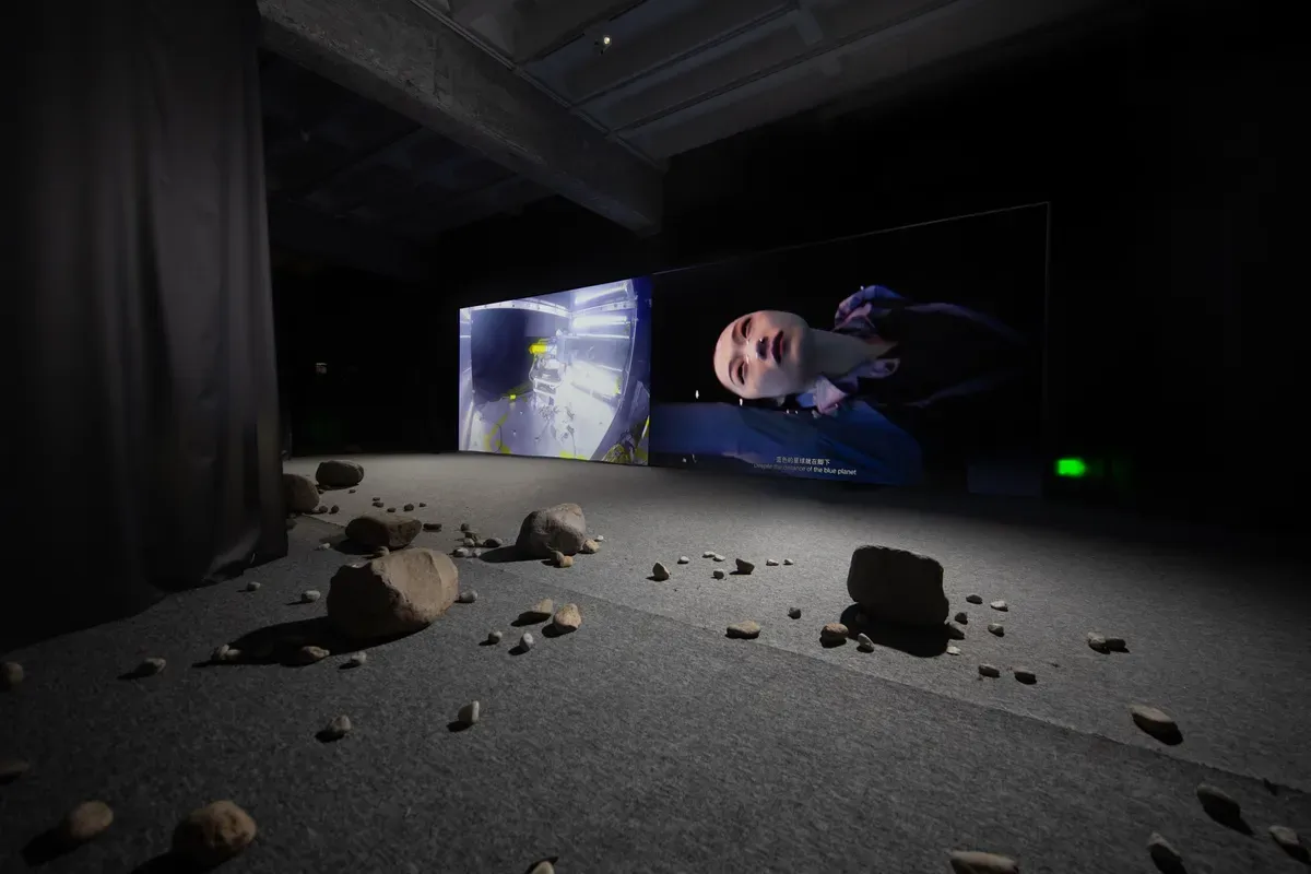 Art installation with rocks scattered on the floor and video displays projecting various images, one of which features a human face.
