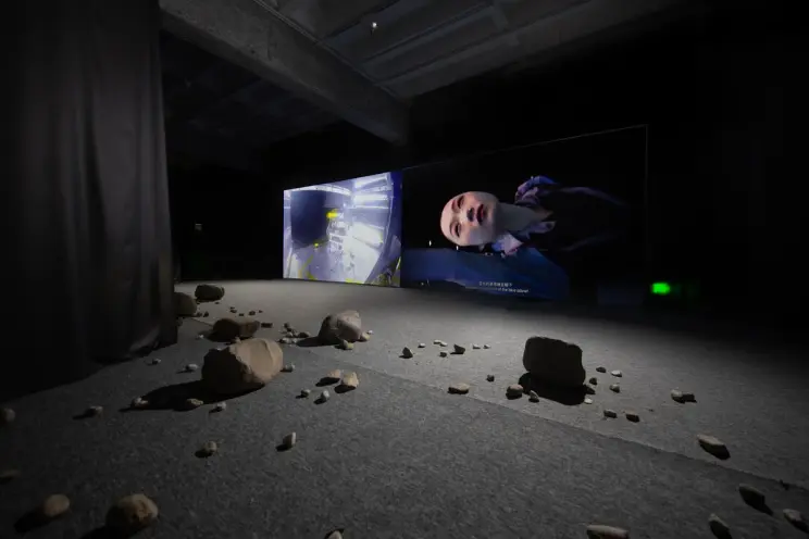 Art installation with rocks scattered on the floor and video displays projecting various images, one of which features a h...