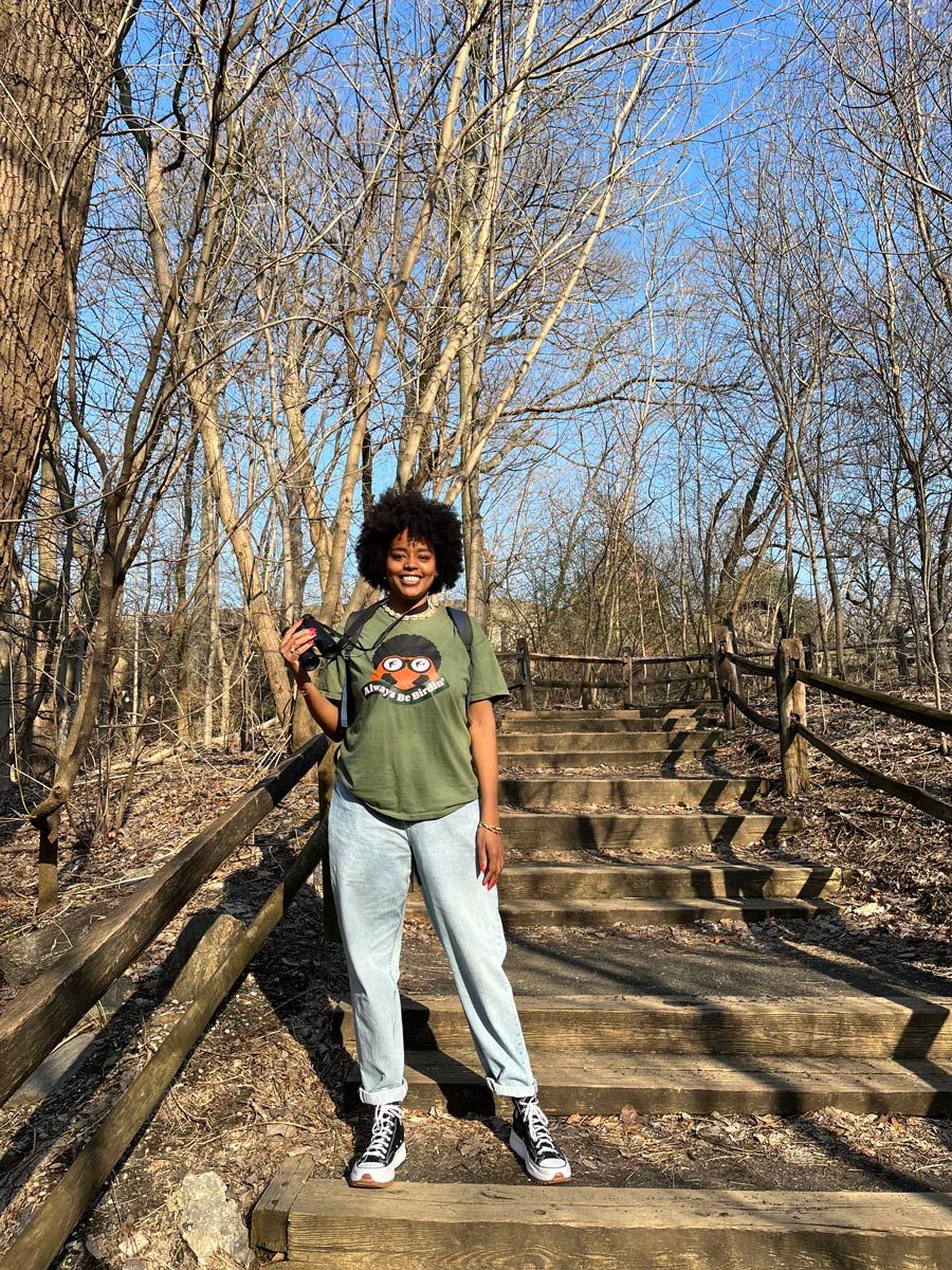 A person standing on a wooden staircase in a forest, holding binoculars and wearing a green shirt with a bird graphic and the words "Always Be Birding."