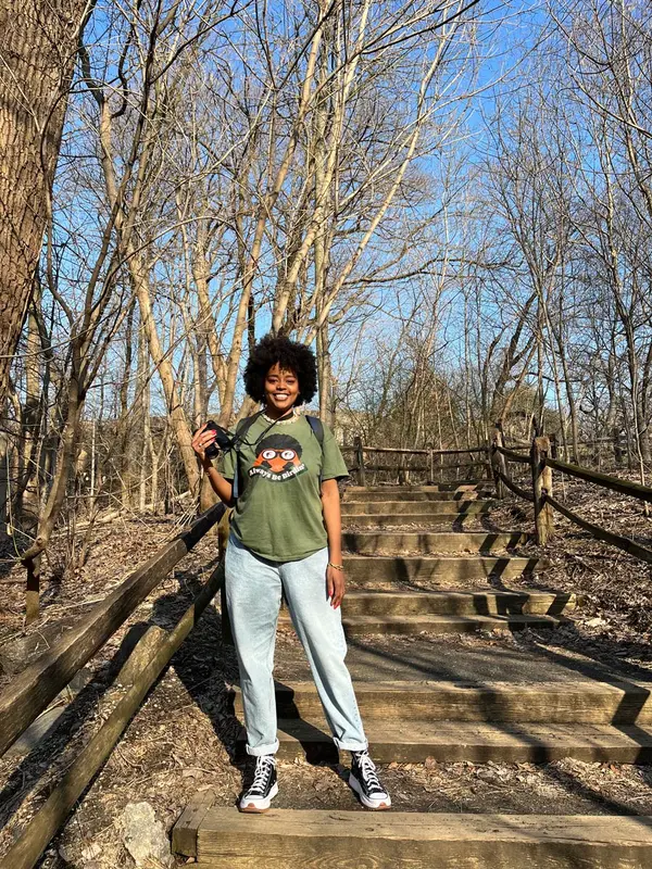 A person standing on a wooden staircase in a forest, holding binoculars and wearing a green shirt with a bird graphic and the words "Always Be Birding."