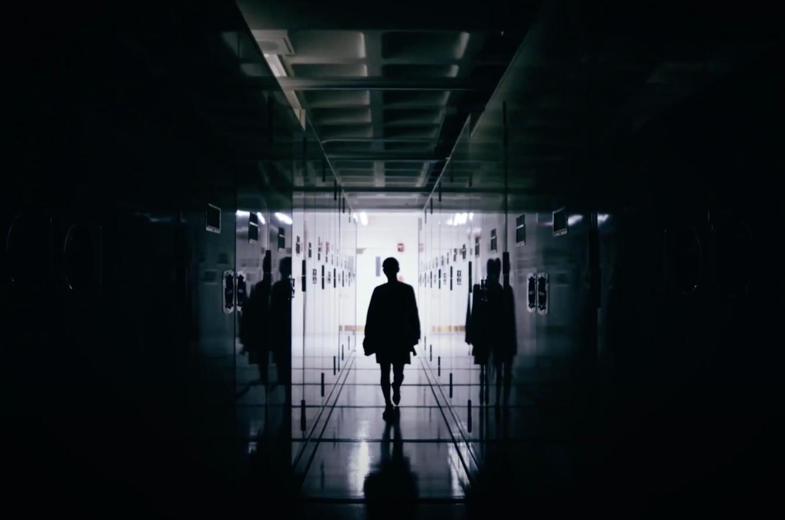 A silhouetted figure strides down a dimly lit, locker-lined hallway, heading towards the radiating light at the corridor's...