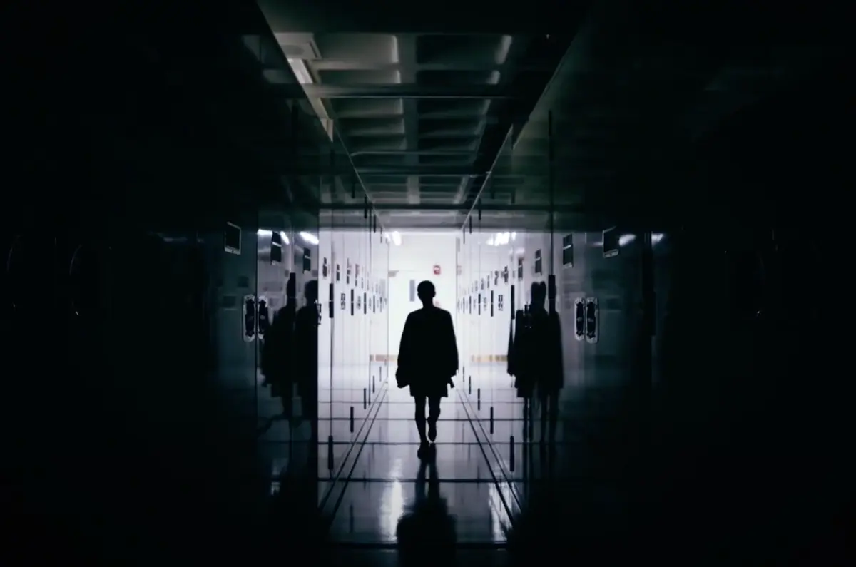 A silhouetted figure strides down a dimly lit, locker-lined hallway, heading towards the radiating light at the corridor's end.