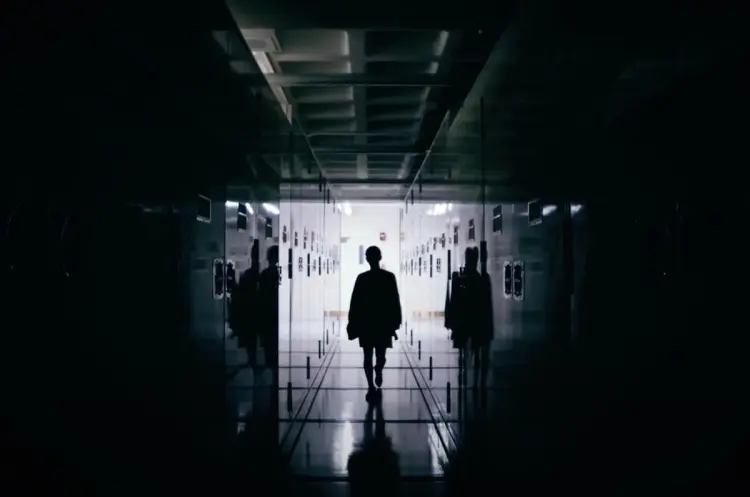 A silhouetted figure strides down a dimly lit, locker-lined hallway, heading towards the radiating light at the corridor's...