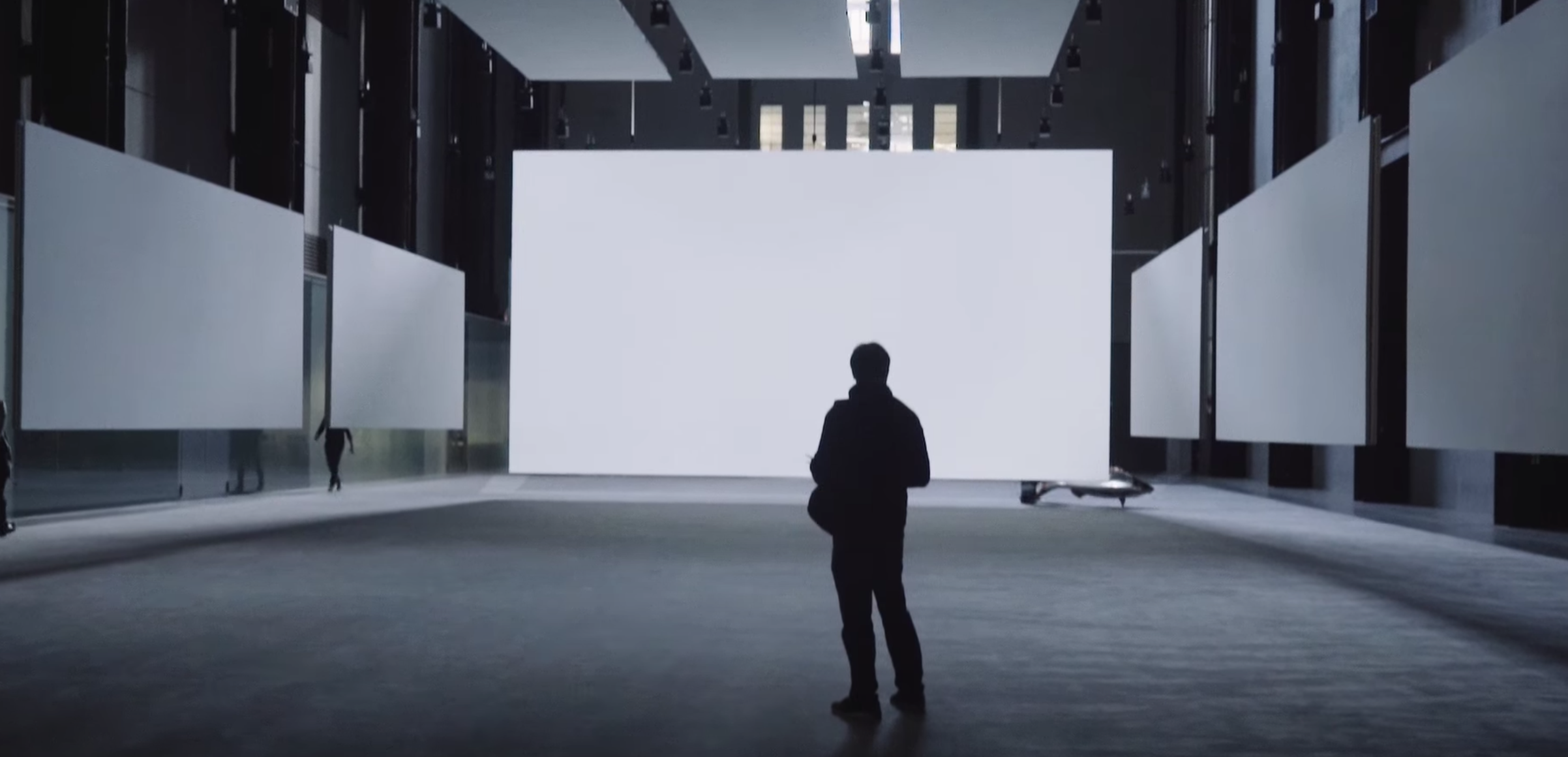 A person standing alone in a spacious gallery hall, encircled by tall white panels. Three hefty, white panels are suspende...