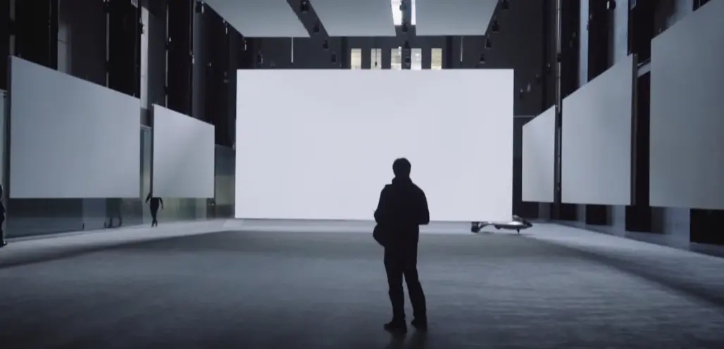 A person standing alone in a spacious gallery hall, encircled by tall white panels. Three hefty, white panels are suspende...
