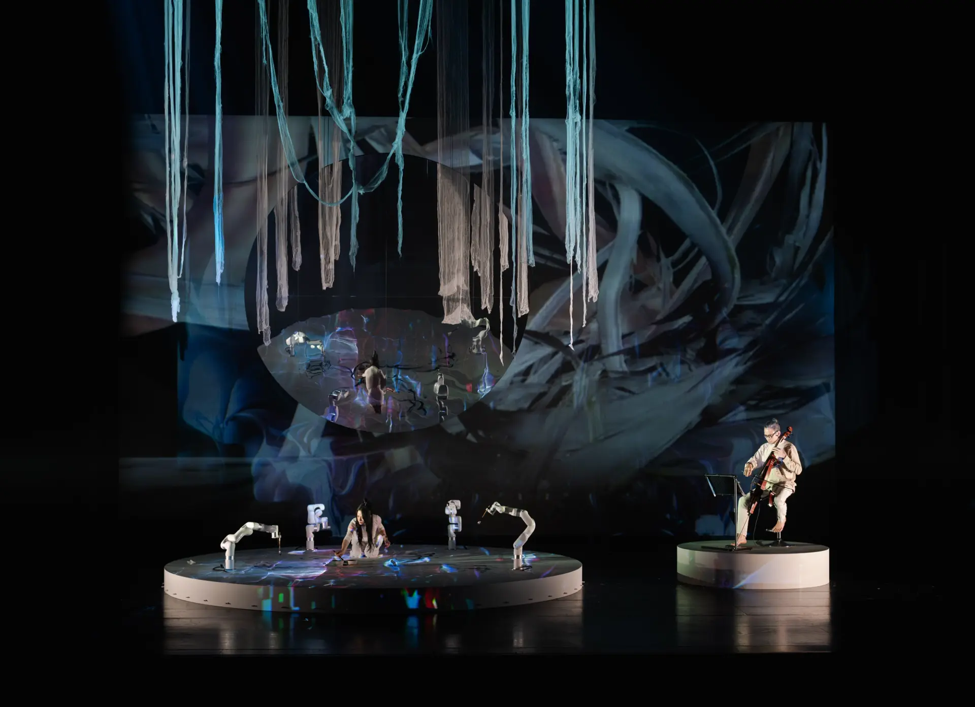 A performer in white interacts with a sheet on the floor under blue lights, reflected in a large round mirror, while another individual plays the cello nearby.