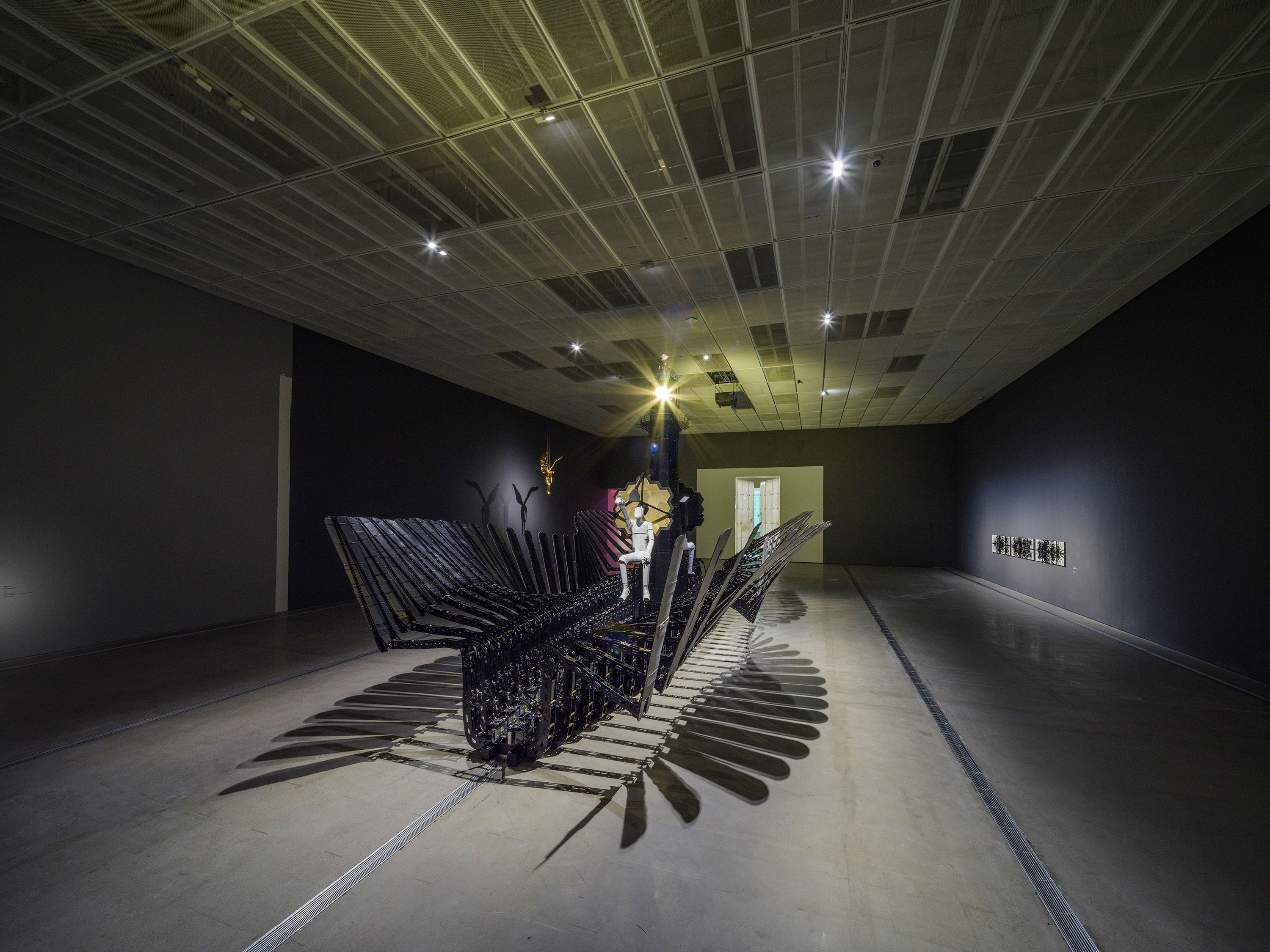 A person is seated atop a large, intricate sculpture made from recycled cardboard boxes and metallic materials. The sculpt...