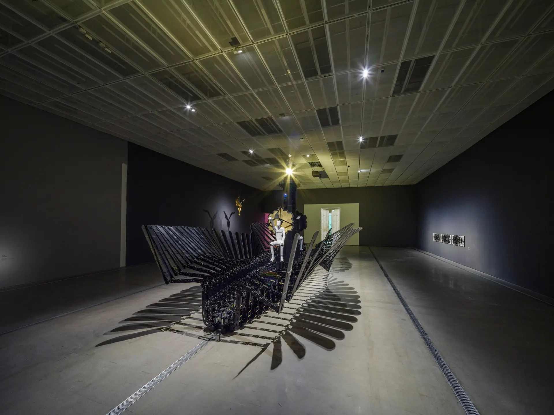 A person is seated atop a large, intricate sculpture made from recycled cardboard boxes and metallic materials. The sculpt...