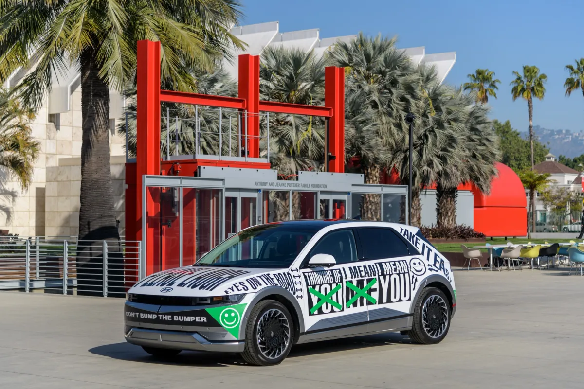 A Hyundai IONIQ 5 vehicle, covered in a digital print vinyl featuring text, is parked in front of a building. The structure has a grey and red facade.