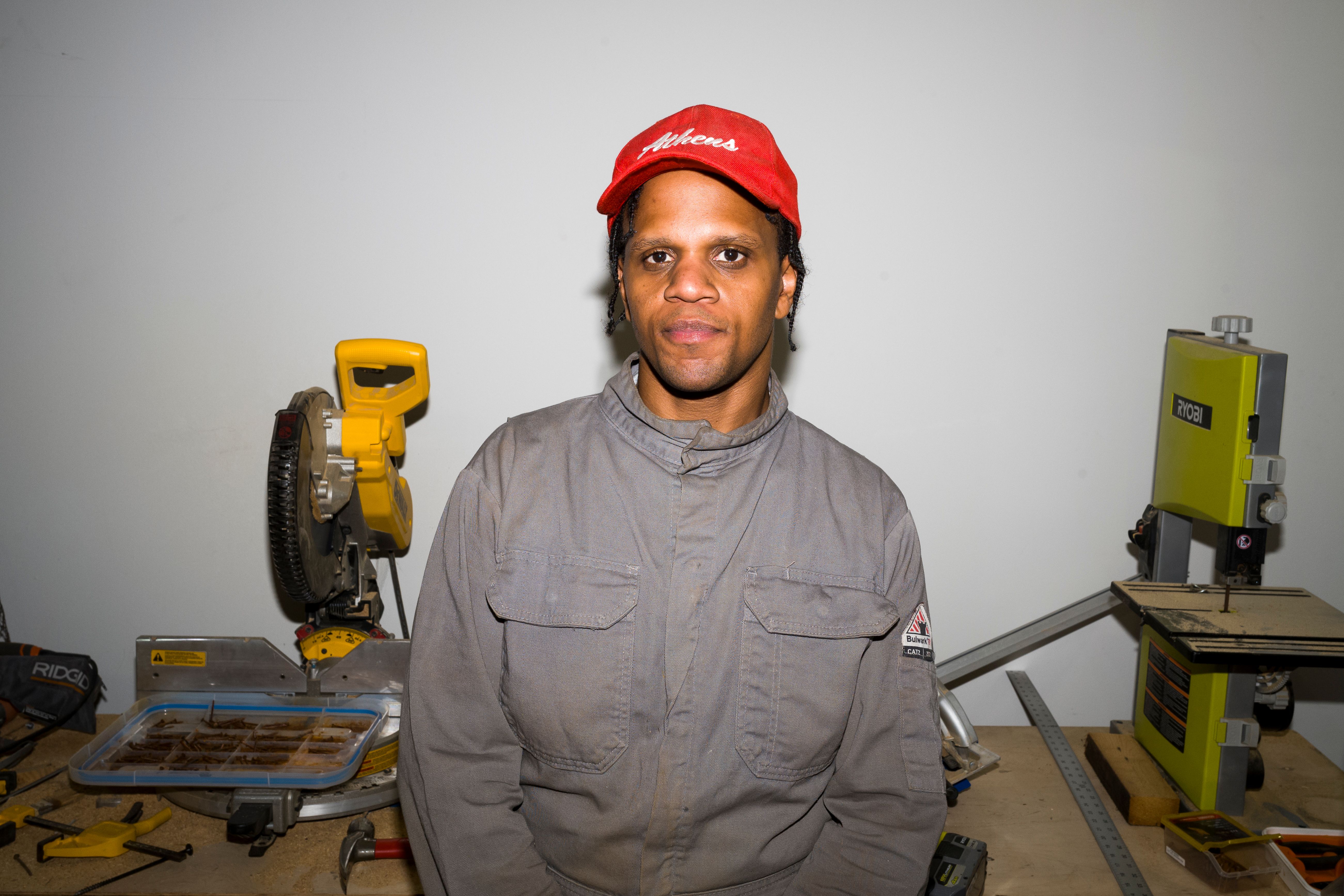 A person in a work suit and red cap stands in a workshop, surrounded by tools and machinery, facing the camera directly.