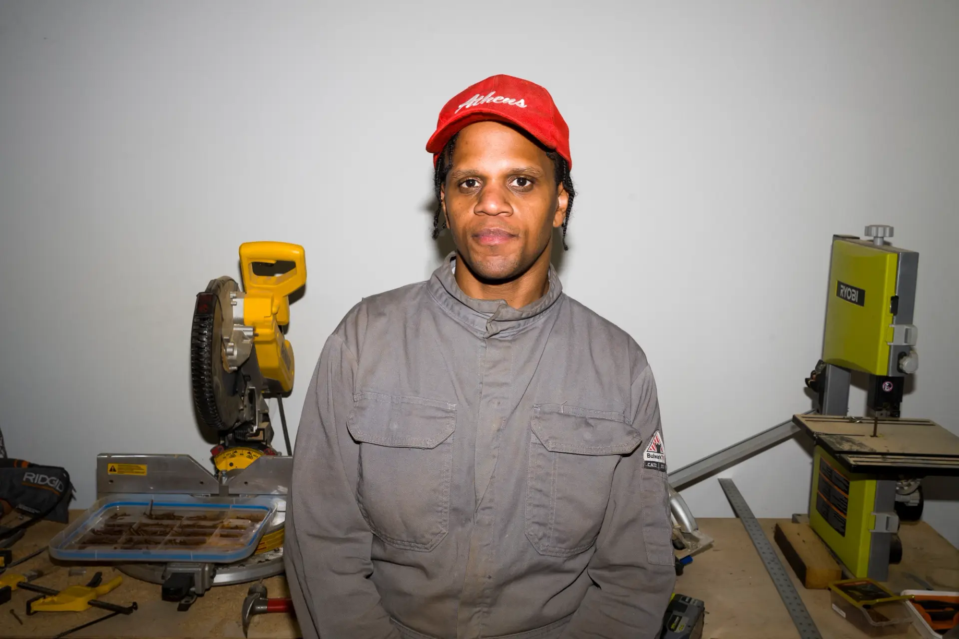 A person in a work suit and red cap stands in a workshop, surrounded by tools and machinery, facing the camera directly.