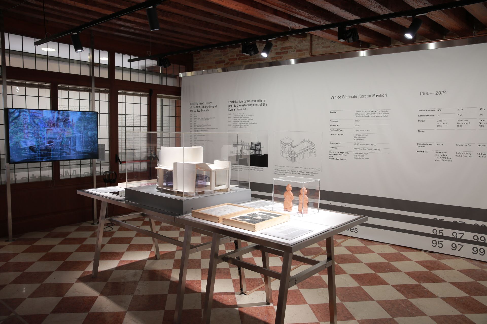 Interior shot of a gallery displaying various objects on tables, such as models and books, against a backdrop of an inform...
