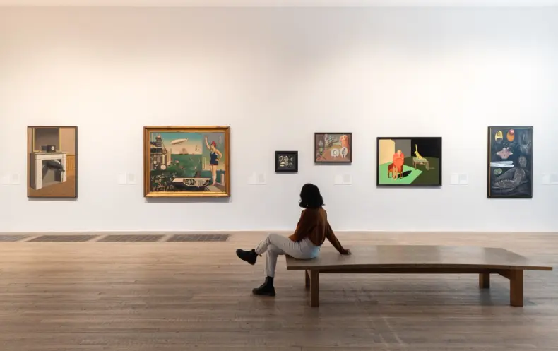 An individual seated on a wooden bench, observing various framed paintings displayed on a gallery wall.