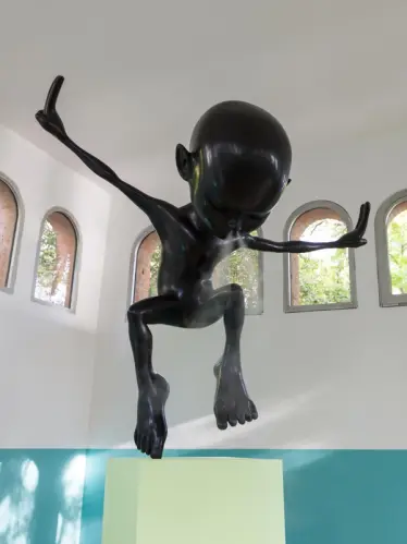 Close-up of a shiny black humanoid sculpture with extended limbs on a green pedestal, set against a white and teal two-ton...