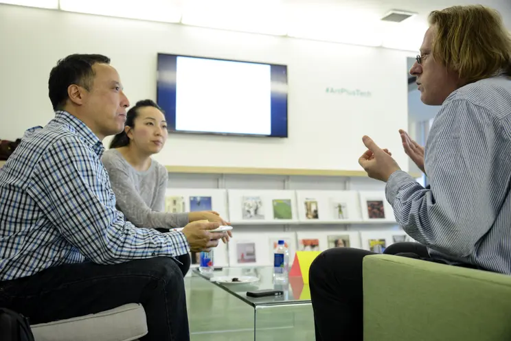 Three individuals, including Jonathon Keats and John Suh, engaged in conversation in a brightly lit room. All are seated c...