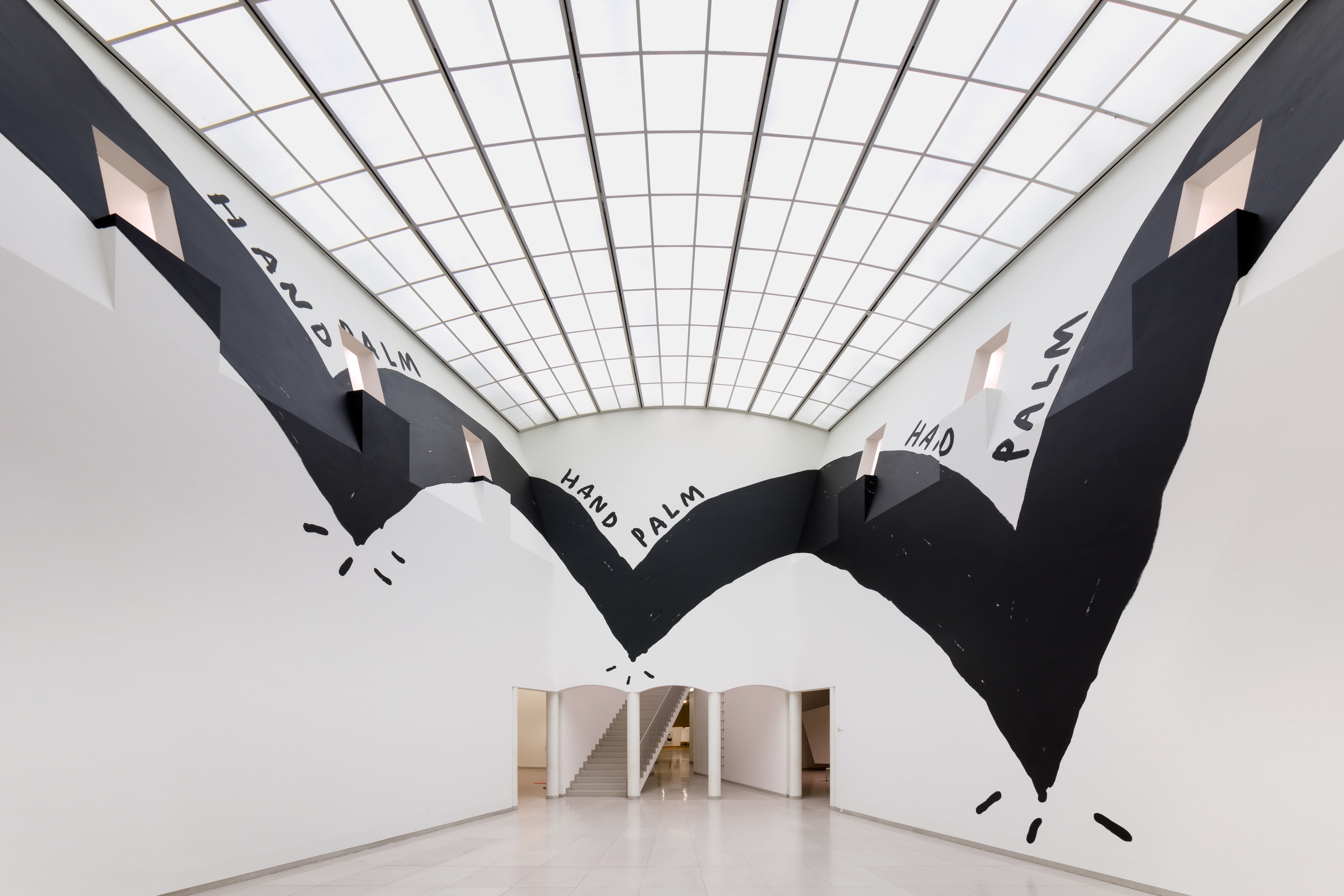 A large-scale wall mural inside a gallery with bold black shapes and the words “HAND PALM HAD PALM”.
