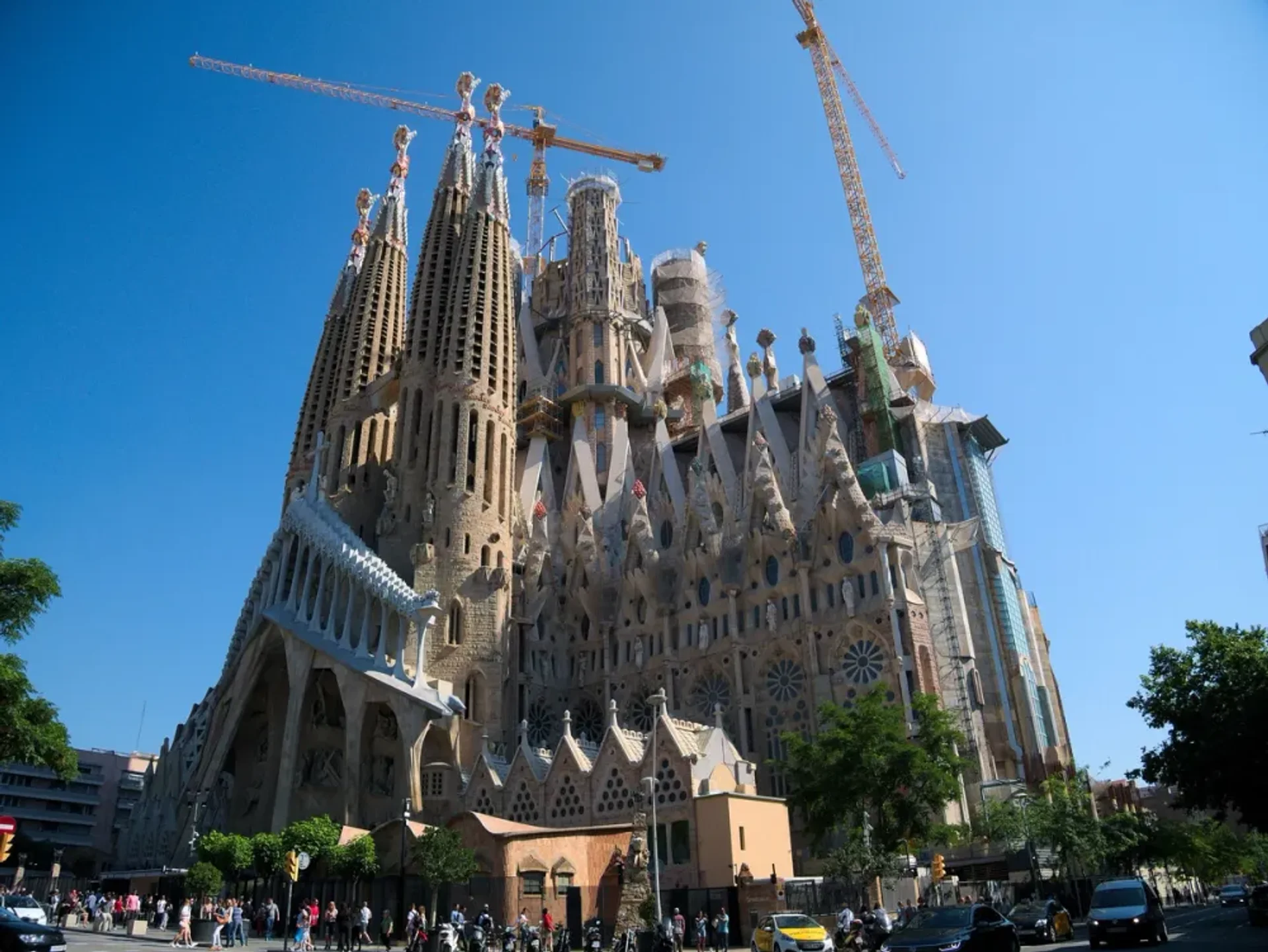 The Gothic and medieval cathedral stands tall against a city skyline, with a towering steeple and intricate design. Construction cranes stand atop the building.