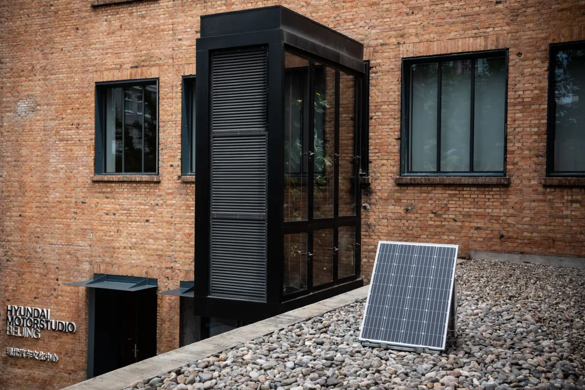 A photovoltaic installation by Tega Brain, titled Solar Protocol, situated outside the Hundai Motorstydio building. The building is constructed of bricks with black windows. Pebbles are scattered on the ground nearby.